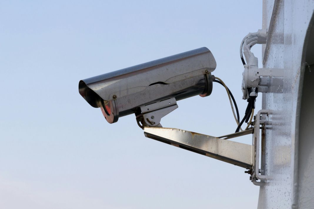 A metallic CCTV camera fixed on the side of a white building, pointing outward.