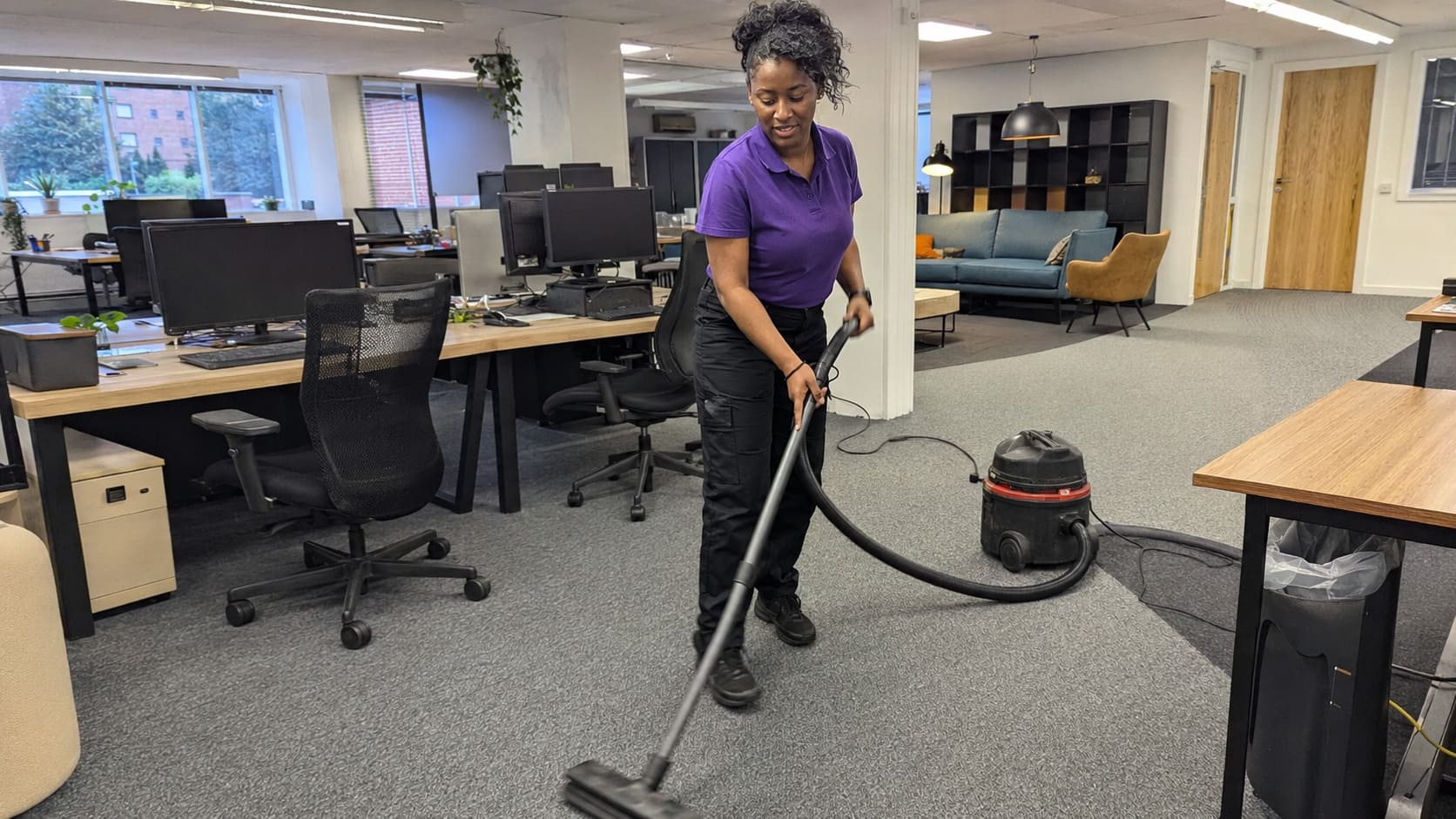 Cleaner vacuuming carpet between desks in a modern open-plan office